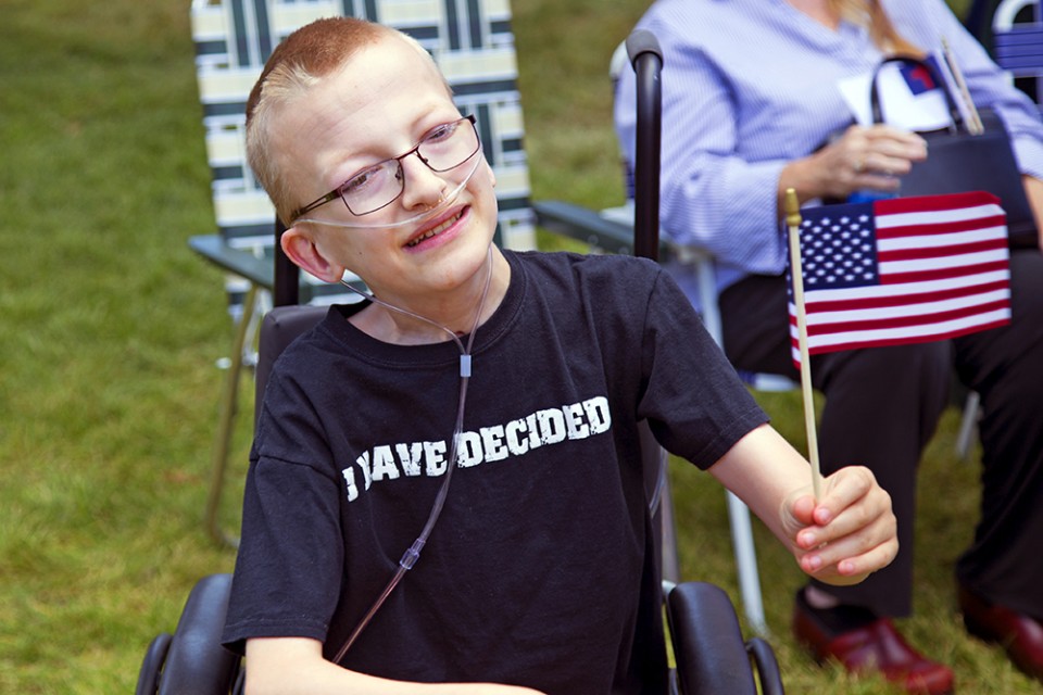 Boy in wheelchair on oxygen, holding American flag and smiling, wearing "I have decided" T-shirt