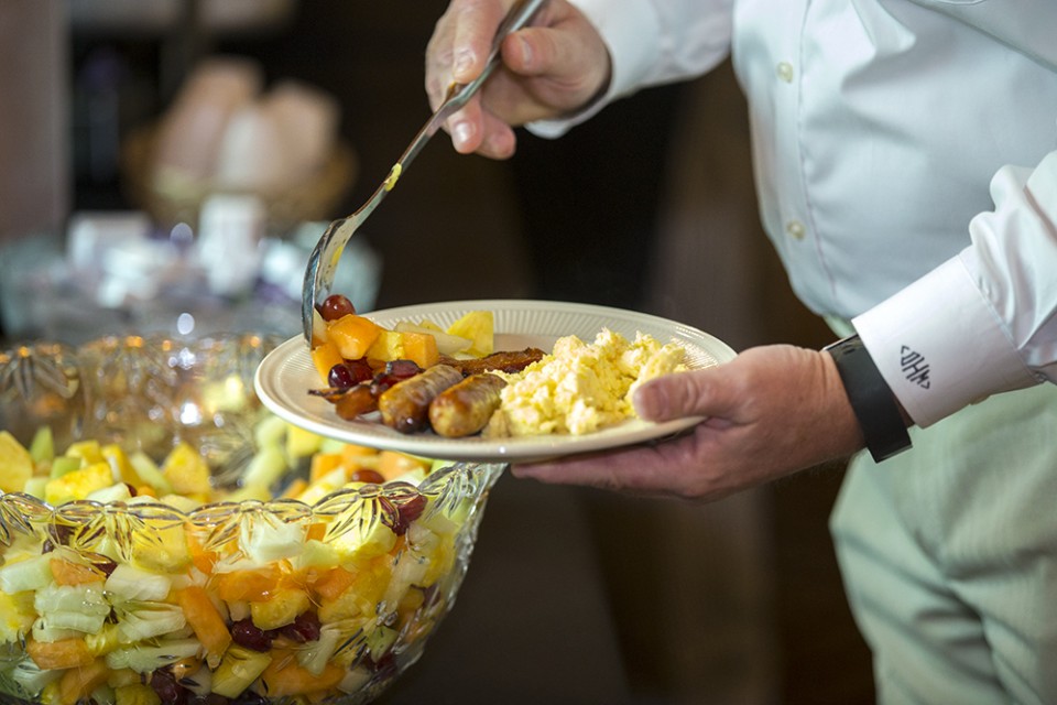 Man's hand scooping fruit on his plate containing sausage and eggs.