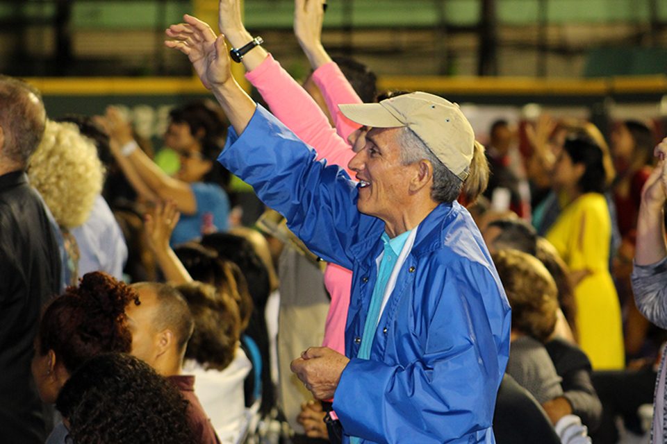 Older man in blue jacket and baseball hat with right arm raised, smiling