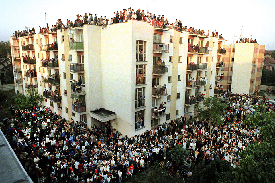 Crowds of people in the street, on balconies and the rooftop of an apartment building.