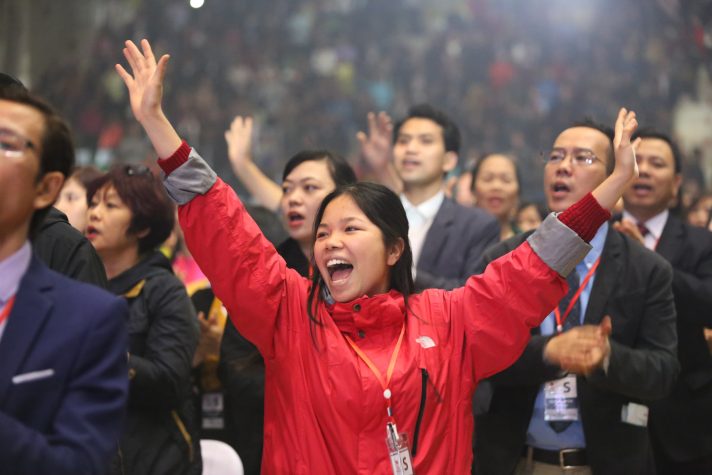 Woman in red jacket, raising hands, singing in the middle of the crowd