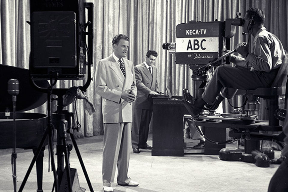 Billy Graham standing in front of an ABC television camera with Cliff Barrows in the background