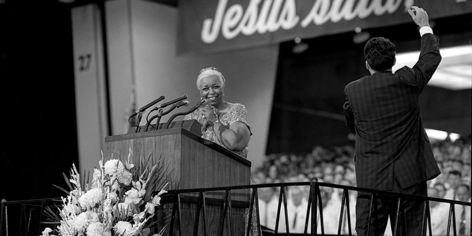 Black and white photo of Ethel Waters at lectern, smiling. Cliff Barrows raises an arm to direct the choir.