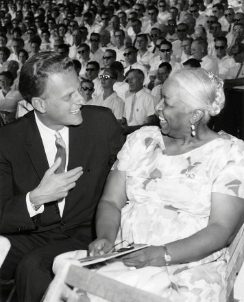 Billy Graham kneels down beside Ethel Waters, smiling.