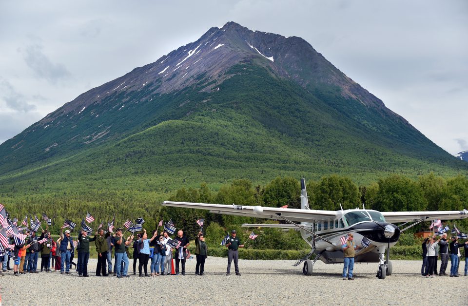 Plane on tarmac with mountain in background, people holding American flags as couples deplane